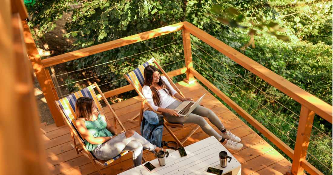 Zwei Frauen sitzen auf Holzbalkon auf Gartenliegen und arbeiten am Laptop